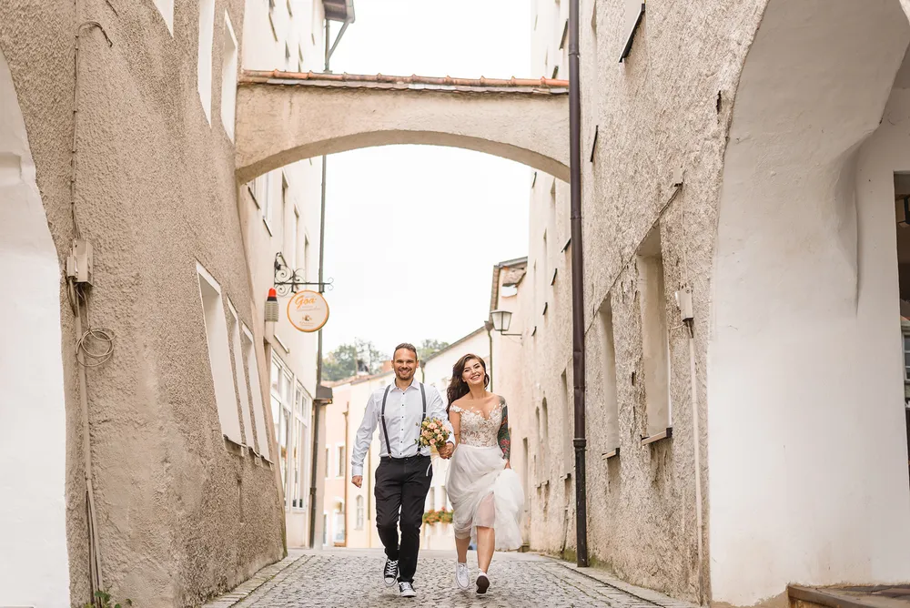 Couple in wedding attire walks down cobblestone street, holding hands, under an arched bridge, surrounded by historic buildings.
