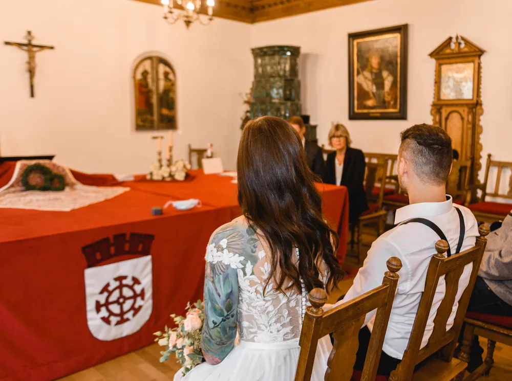 Bride in lace wedding dress with floral tattoos, holding bouquet, seated in church aisle. Groom in white shirt with suspenders, facing altar with red cloth and coat of arms. Clergy and guests seated at the altar.