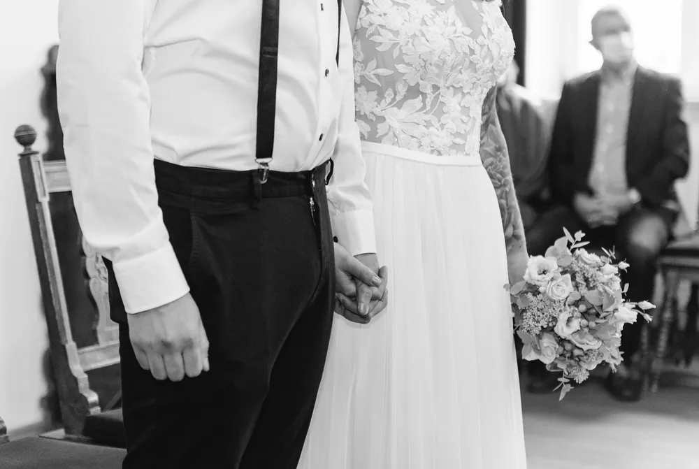 A bride in a lace gown and a groom in a classic suit hold hands at a wedding ceremony, with a guest in the background.