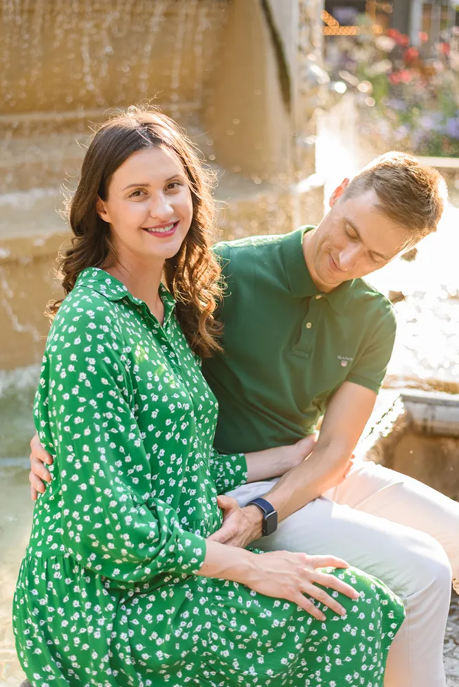A couple sits closely by a fountain; the woman in a green floral dress smiles, the man in a green polo leans in, both hands clasped, against a backdrop of blurred water and foliage.