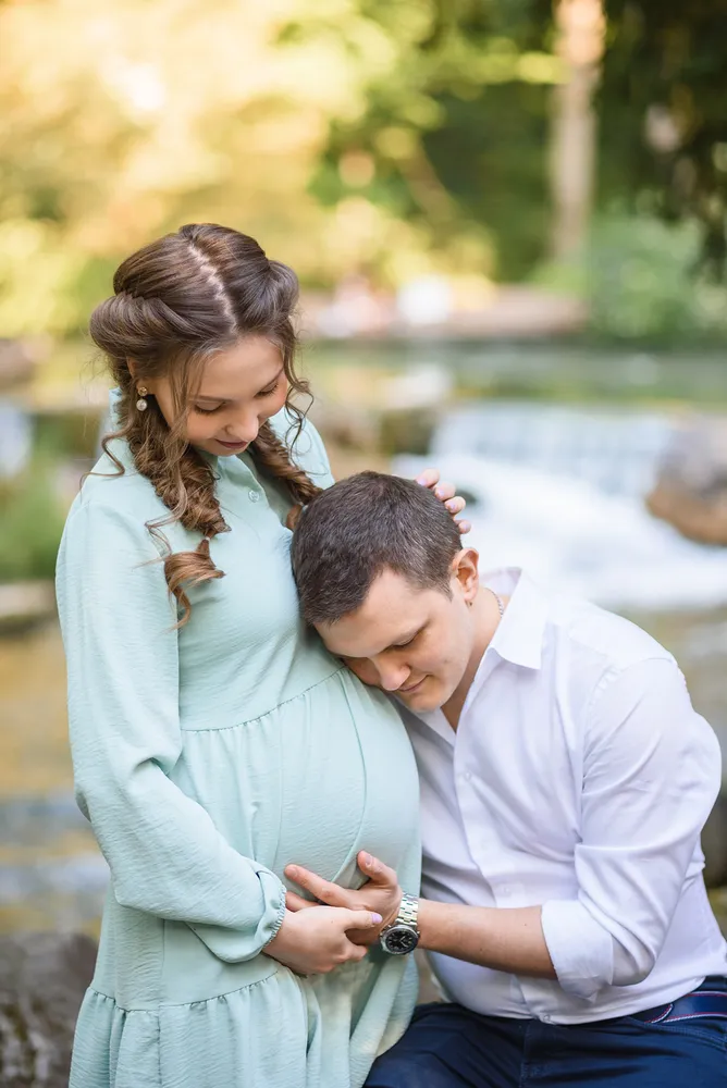 Couple embracing outdoors; woman in light green dress cradles belly, man in white shirt kneels beside her, both smiling softly, serene natural backdrop.