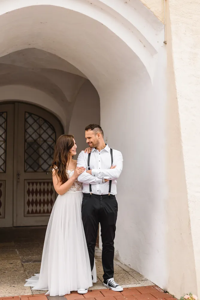 A couple in wedding attire stands under an archway, smiling and embracing, with a decorative door behind them.