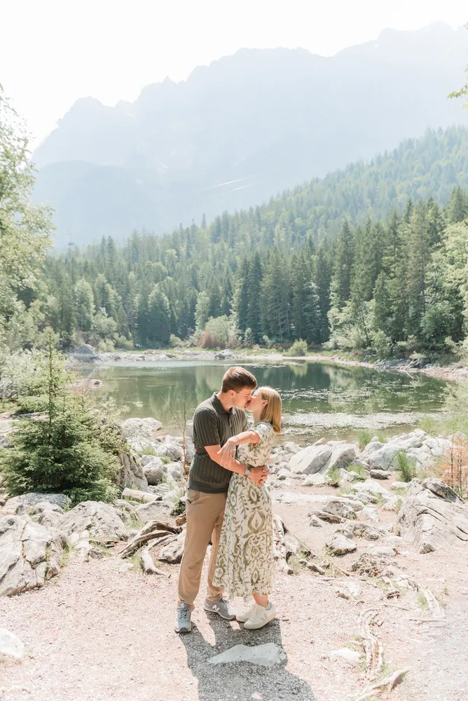 Couple embracing by serene lake, surrounded by forest and mountains. Man in striped shirt, woman in floral dress. Peaceful outdoor setting.
