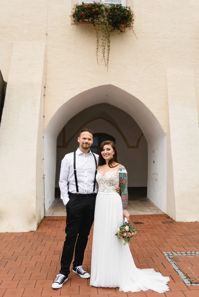 A couple stands smiling before a stone archway, the bride in a white gown with floral tattoos, holding a bouquet, the groom in a white shirt with black suspenders and sneakers.