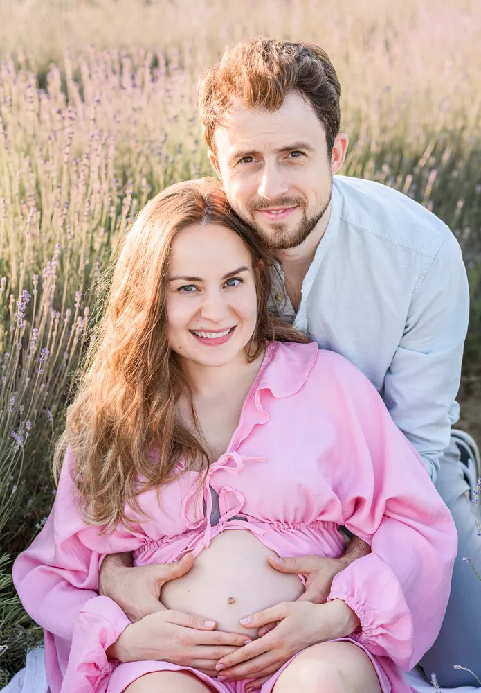 Expectant couple in a lavender field. Woman in pink blouse cradles her belly. Man in light blue shirt behind her, both smiling. Natural daylight, soft focus background.