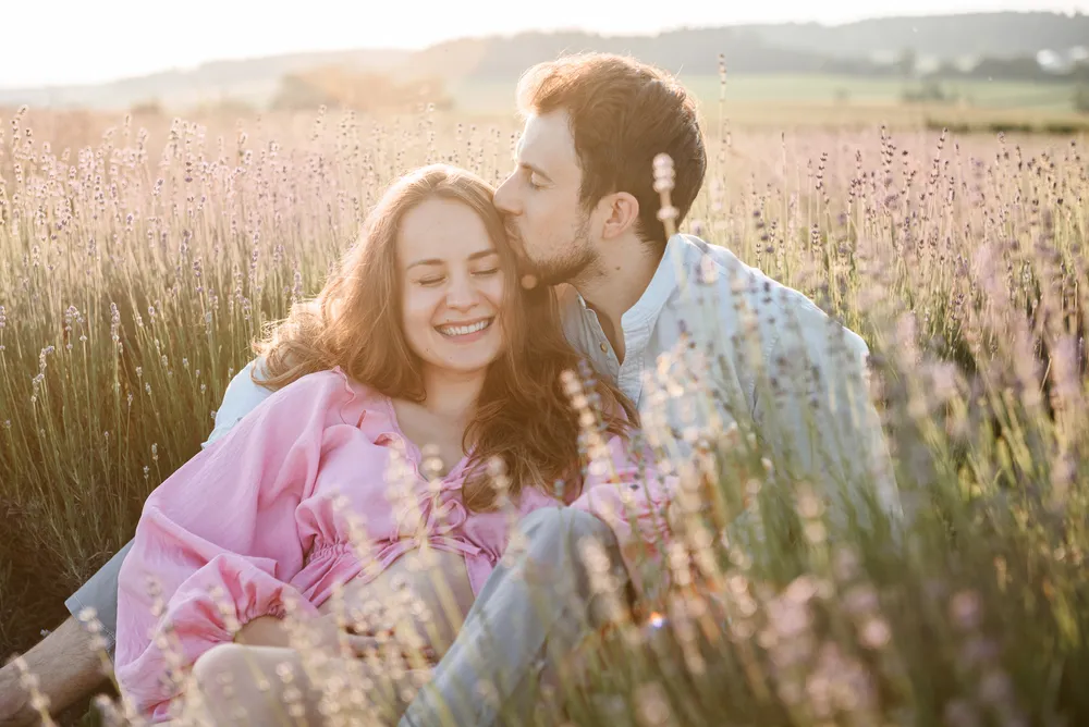 Couple shares an affectionate moment in a lavender field at sunset. Woman in pink dress smiles, man leans in for a kiss. Warm, golden light bathes the scene.
