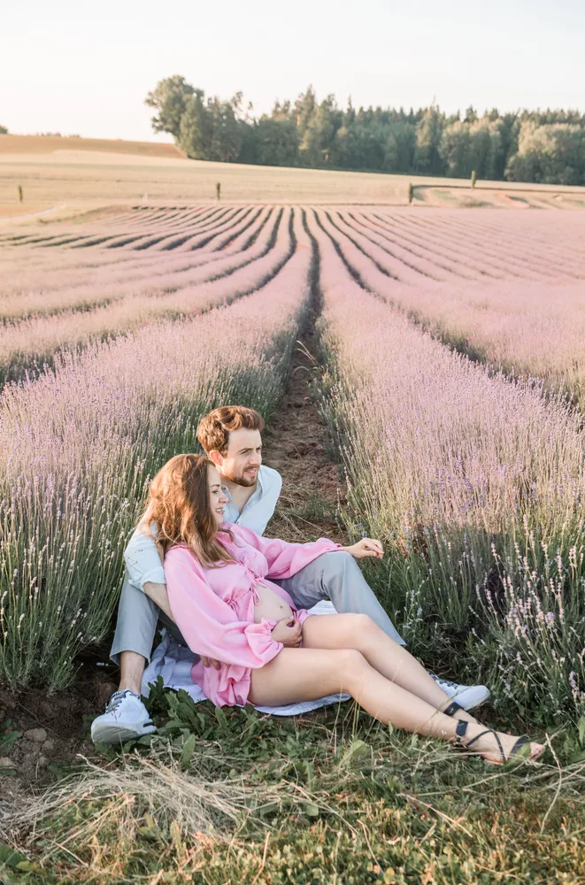 Couple seated in a lavender field, man in white shirt, woman in pink dress, surrounded by purple flowers under a clear sky.