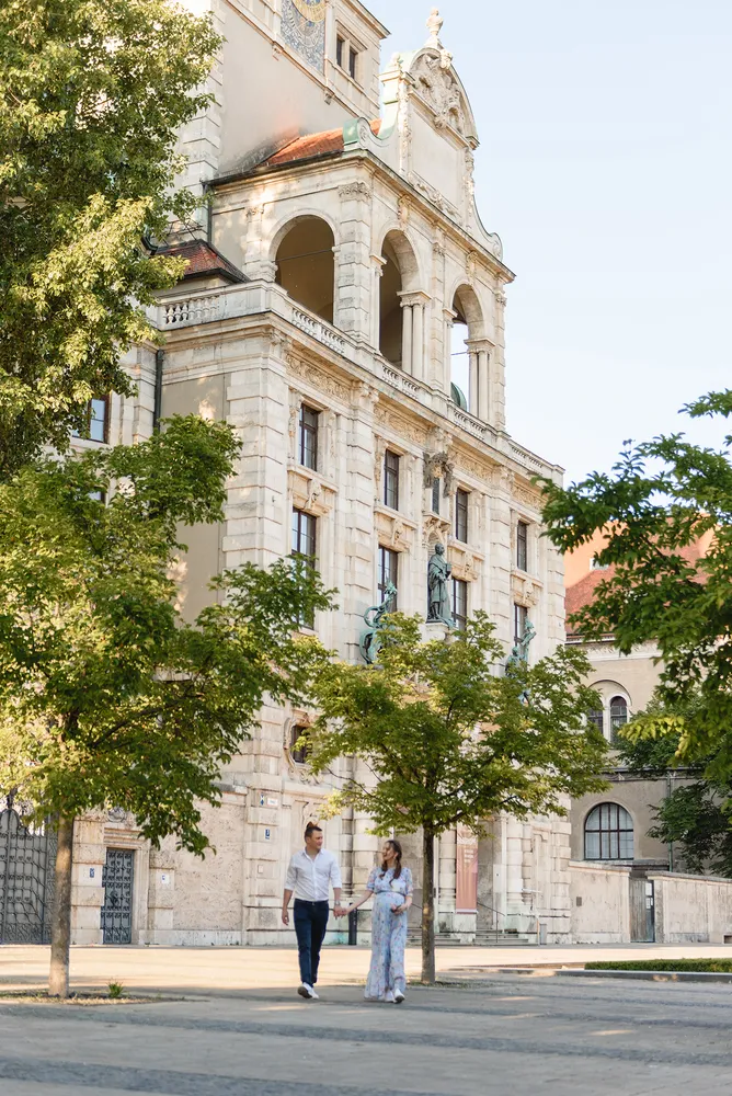 Couple strolls on cobblestone street beside ornate historical building with trees and statues.