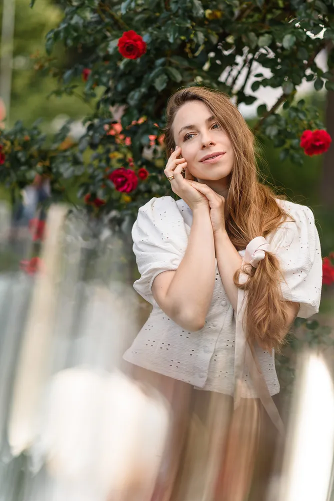 Woman in white blouse with lace details stands outdoors, hand resting on face, amidst a backdrop of red roses and greenery. Soft focus on surroundings.