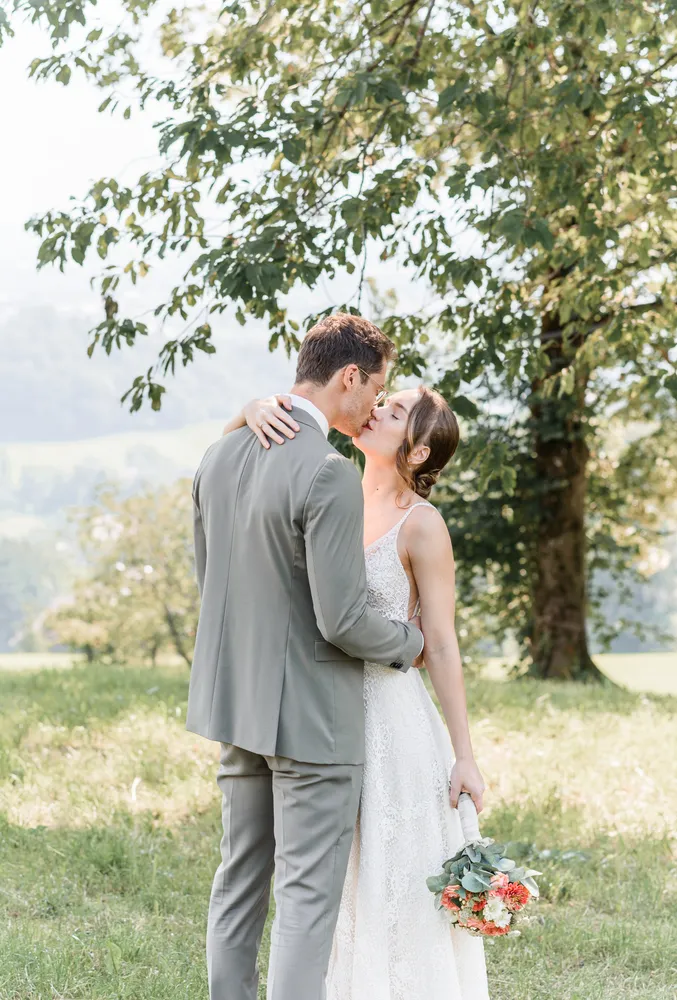 A couple in wedding attire shares a kiss outdoors, she in a lace dress holding a bouquet, he in a gray suit, under a tree with a scenic backdrop.