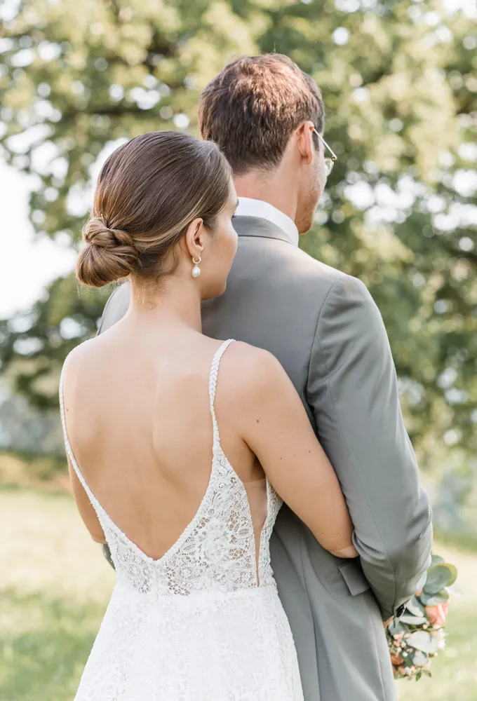 Bride in lace wedding dress, groom in gray suit, holding hands, standing in park with trees and grass, looking at something in distance.