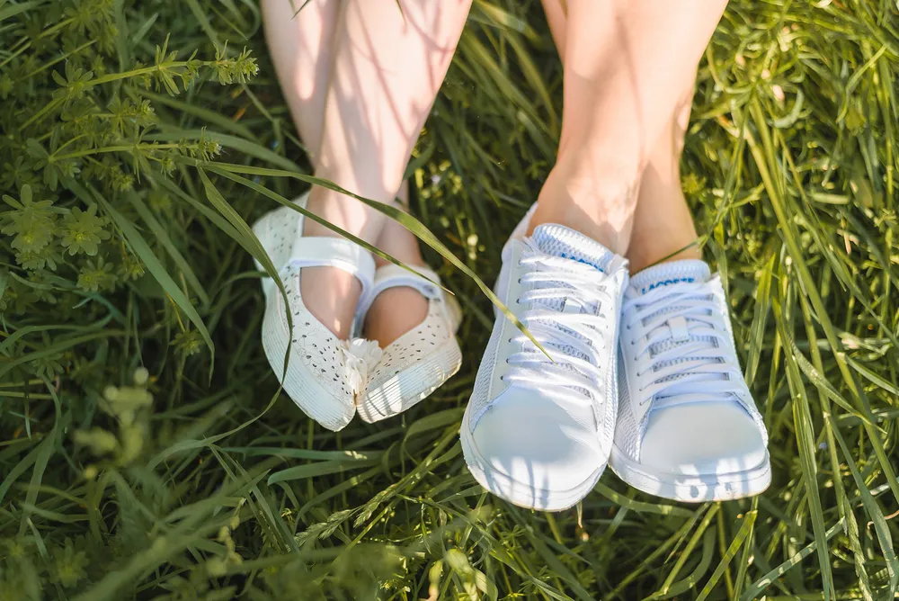 Two pairs of feet, one in white lace-up sneakers and the other in white perforated sandals, rest on lush green grass. The scene is sunlit, highlighting the contrast between the footwear and the verdant surroundings.