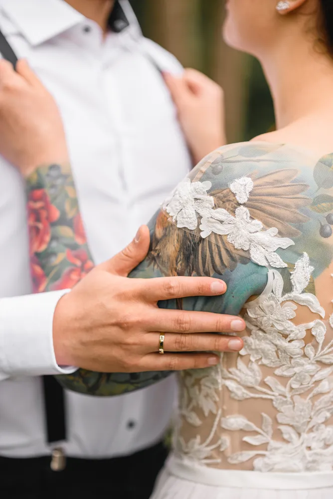 A couple in wedding attire stands close; the groom's tattooed arm rests on the bride's lace-detailed back, their hands gently clasped, hinting at a shared moment in a softly focused outdoor setting.
