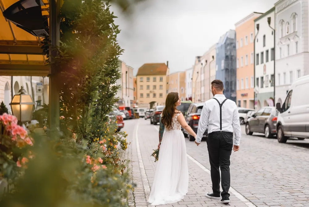 Couple in wedding attire strolls down cobblestone street, holding hands, urban backdrop with parked cars and European architecture.