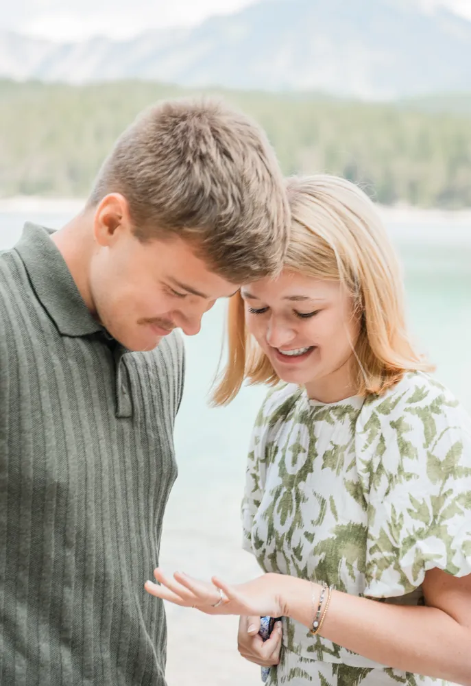 A young couple stands by a serene lake, smiling as they share a joyful moment. The man in a grey sweater and the woman in a floral dress are the focal points of this outdoor photograph, with a picturesque mountain range softly blurred in the background.