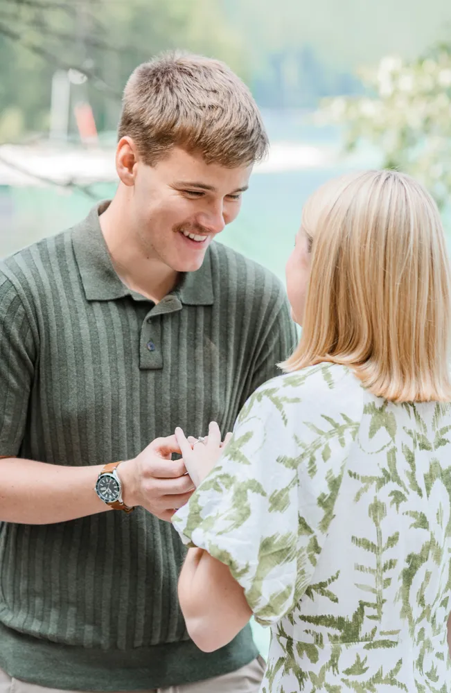 Man and woman stand close, smiling, he holds her hand, she gazes at ring. Green polo, floral blouse. Outdoors, blurred nature background.