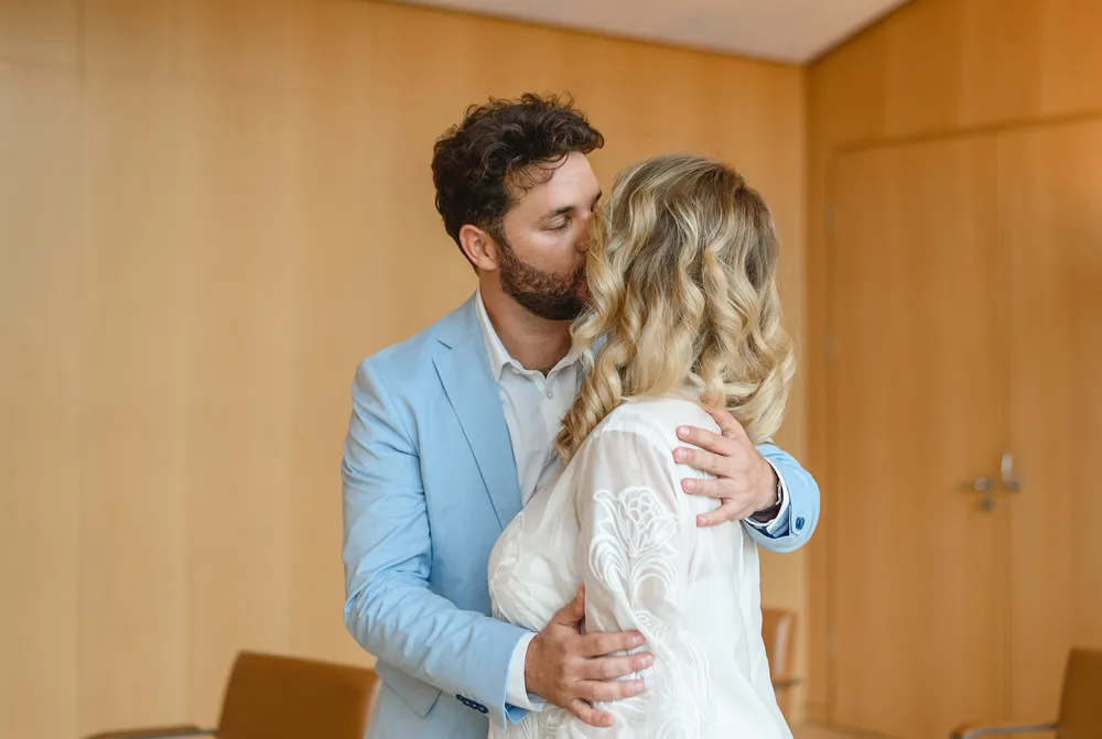 Man in blue suit kisses woman in white dress. They embrace in a room with wooden walls and chairs.