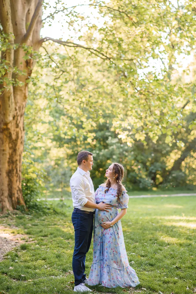 Expectant couple shares a tender moment in a sunlit park, her hand resting on her belly, his support beside her, surrounded by verdant greenery and a large tree.