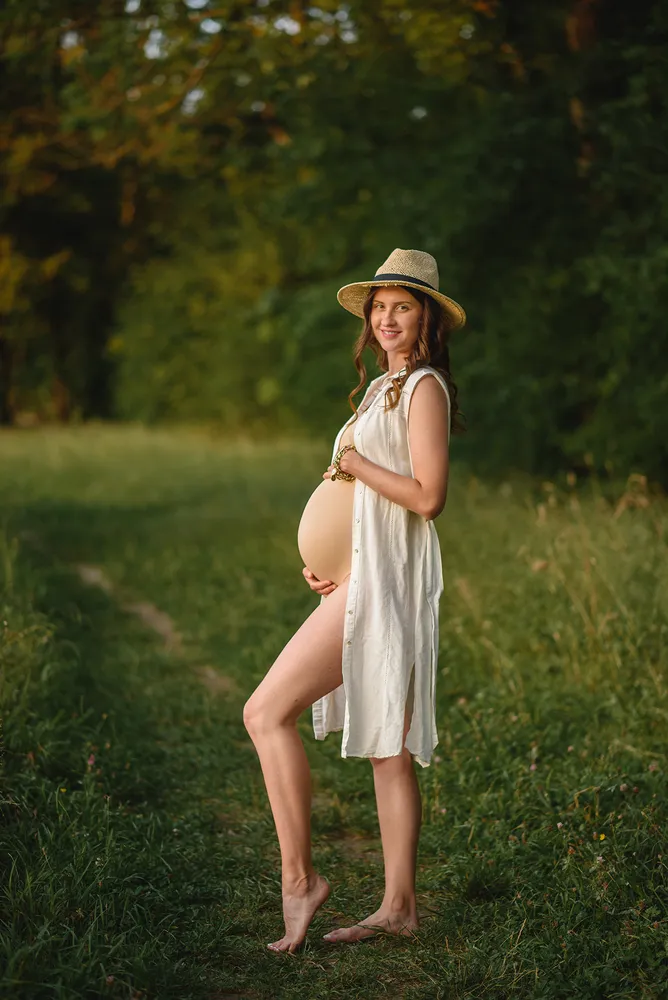 Pregnant woman standing barefoot in a grassy field, wearing a white dress and hat, gently cradling her belly, surrounded by greenery, natural sunlight.
