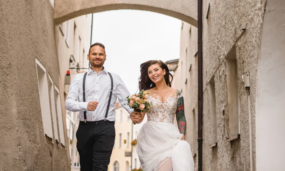 Newlyweds joyfully walk hand-in-hand down a historic alleyway. The groom in a white shirt and suspenders, the bride in a lace-trimmed gown holding a bouquet. Their smiles and relaxed attire suggest a post-ceremony celebration. The narrow passage, flanked by old stone buildings, adds a timeless charm to the scene.