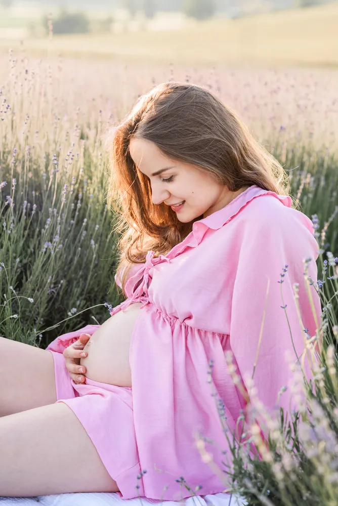 Expectant mother in pink dress caresses belly amidst blooming lavender field at golden hour.