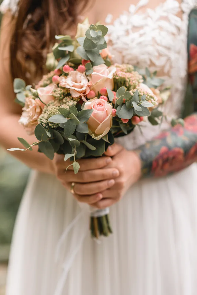 Bride holding a bouquet of peach roses and greenery. She wears a white lace dress with a floral tattoo on her arm. The bouquet is tied with a white ribbon. The background is blurred, focusing on the bouquet and the bride's hands.