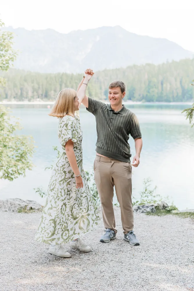 Couple dancing by a lake, woman in floral dress, man in striped polo, joyful expressions, mountainous backdrop, gravel path, clear sky.
