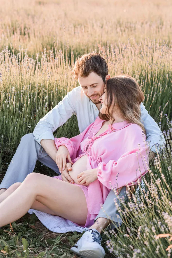 Expectant couple embraces in lavender field. Man supports pregnant woman's belly. Both wear light pink shirts. Serene, natural setting with tall purple flowers. Soft sunlight enhances intimate moment.