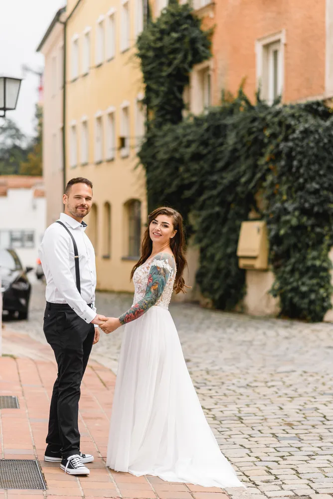 Couple in wedding attire stands on cobblestone street, holding hands. Man wears white shirt, black pants, and sneakers. Woman in white lace wedding dress with floral tattoo visible. Background features ivy-covered buildings.