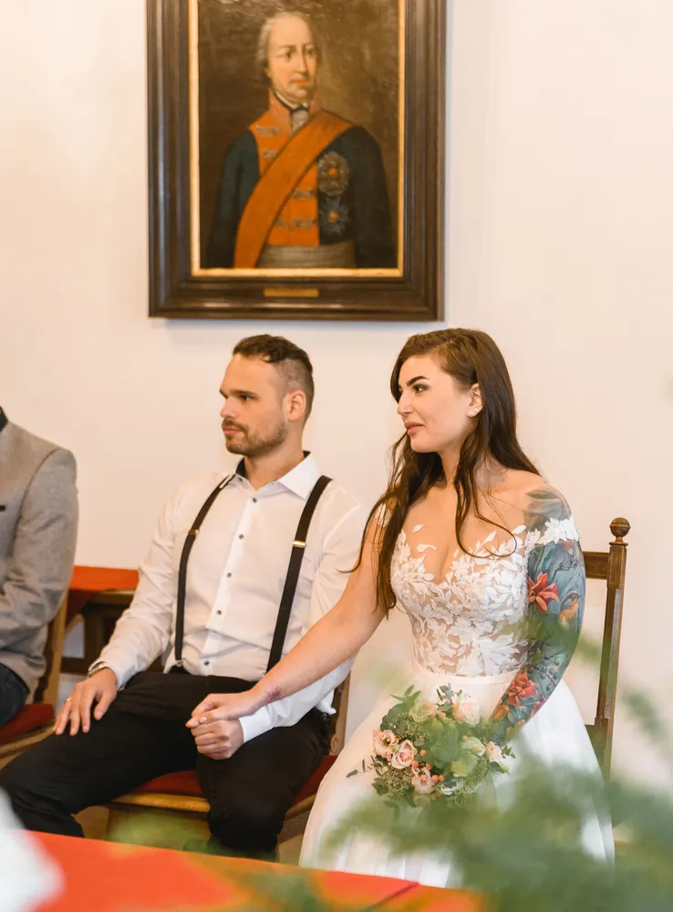 Bride and groom seated at wedding ceremony. Bride with floral tattoo holds bouquet. Groom in white shirt and suspenders. Historical portrait in background. Formal setting.