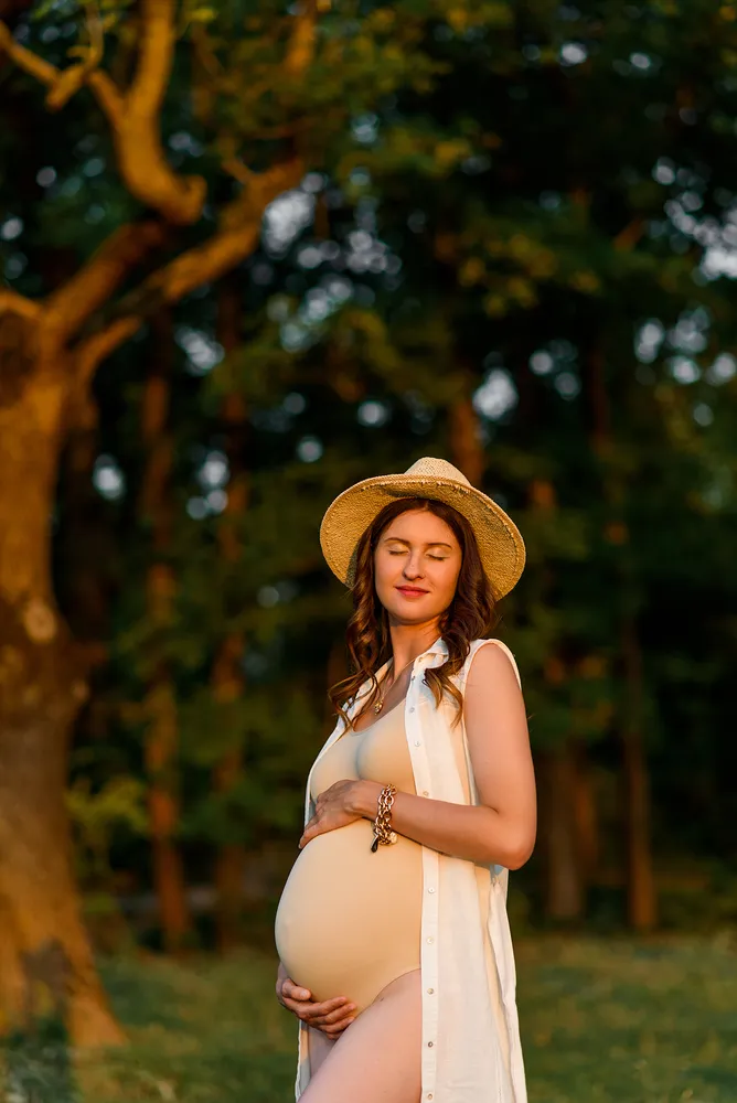 Pregnant woman stands in a forest, gently cradling her belly. She wears a straw hat, white sleeveless top, and gold bracelet. Soft, natural light filters through the trees, creating a serene and intimate atmosphere.