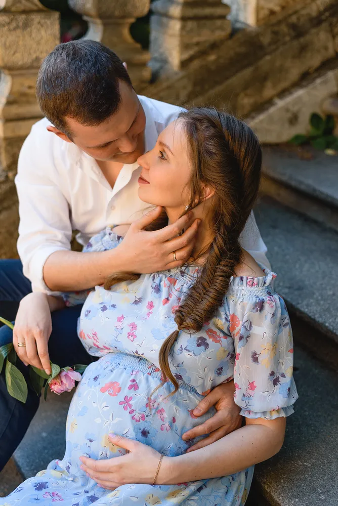 Couple embracing outdoors, man in white shirt holding rose, woman in floral dress cradling visible baby bump, stone steps and greenery in background.