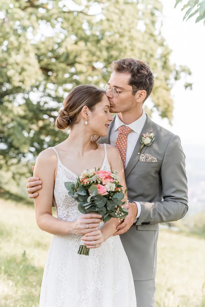 Bride and groom embrace outdoors; he kisses her forehead. She holds pink and white bouquet. He wears grey suit with patterned pocket square. She wears white lace dress. Background: blurred greenery and trees.