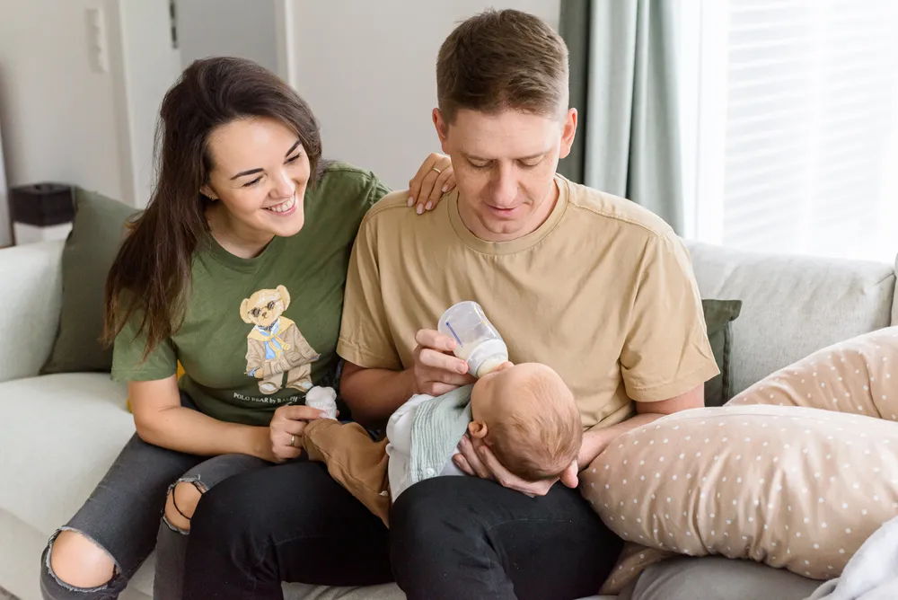 A family of three sits on a couch. A man feeds a baby with a bottle while a woman smiles beside them. Casual clothing, home interior, daylight.