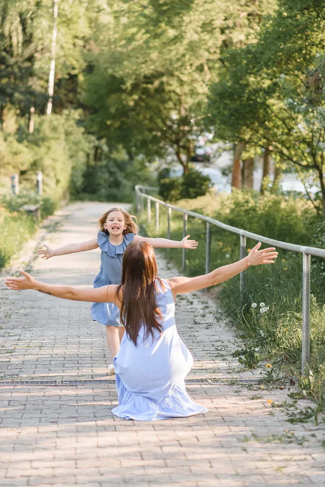 A woman and a young girl in matching blue dresses joyfully embrace on a sunny pathway, surrounded by lush greenery and a gentle river in the background, both with arms wide open in a moment of shared happiness.