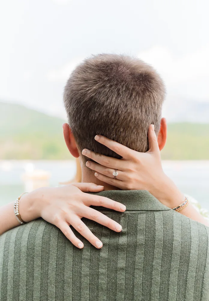Woman's hands tenderly embrace man's neck, diamond ring gleaming, as they stand before serene landscape.