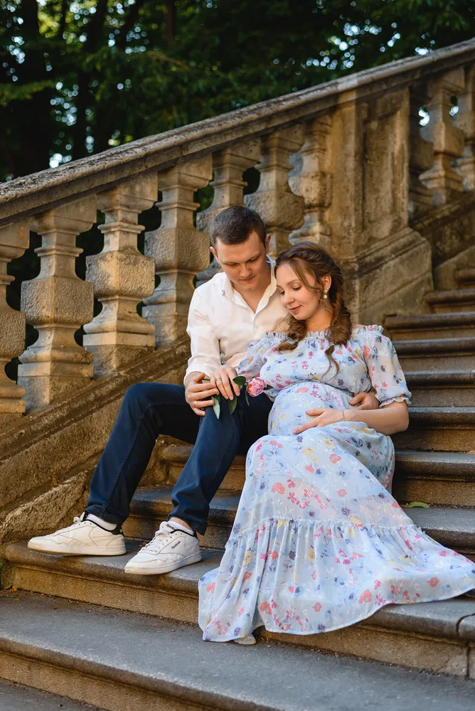 Couple seated on stone steps, woman in floral dress cradles baby bump, man in white shirt holds pink rose, classical balustrade, lush greenery backdrop, serene moment captured.