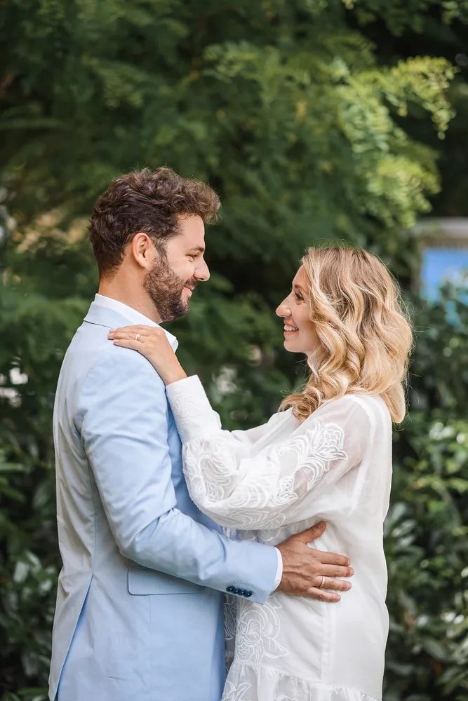 Couple in wedding attire embracing outdoors, man in light blue suit, woman in white lace dress, surrounded by greenery.