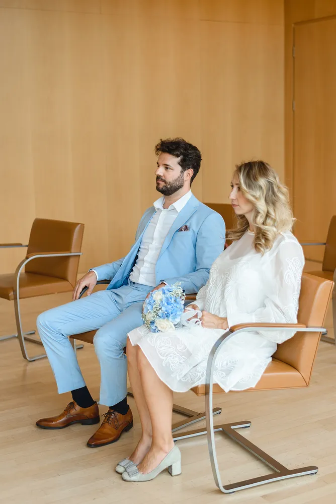 A couple in wedding attire sits in a modern room. The man wears a blue suit, the woman a white lace dress. They hold hands, gazing forward. Wooden walls and chairs surround them. A bouquet rests in the woman's lap.
