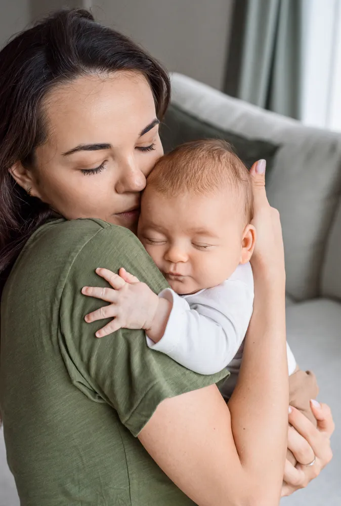 A woman in a green shirt embraces a sleeping baby in white clothing, both with closed eyes, in a softly lit room with a gray couch and curtains in the background.