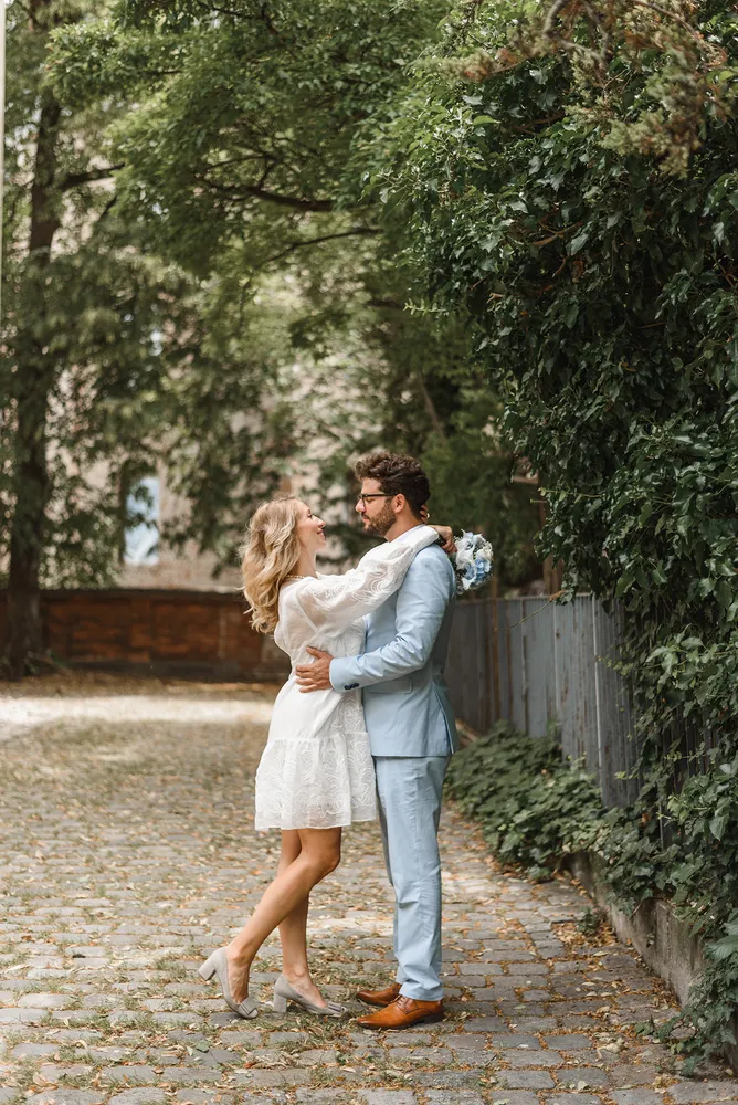 Couple in wedding attire embracing on cobblestone path, surrounded by lush greenery and a rustic fence, with a blurred historic building in the background.