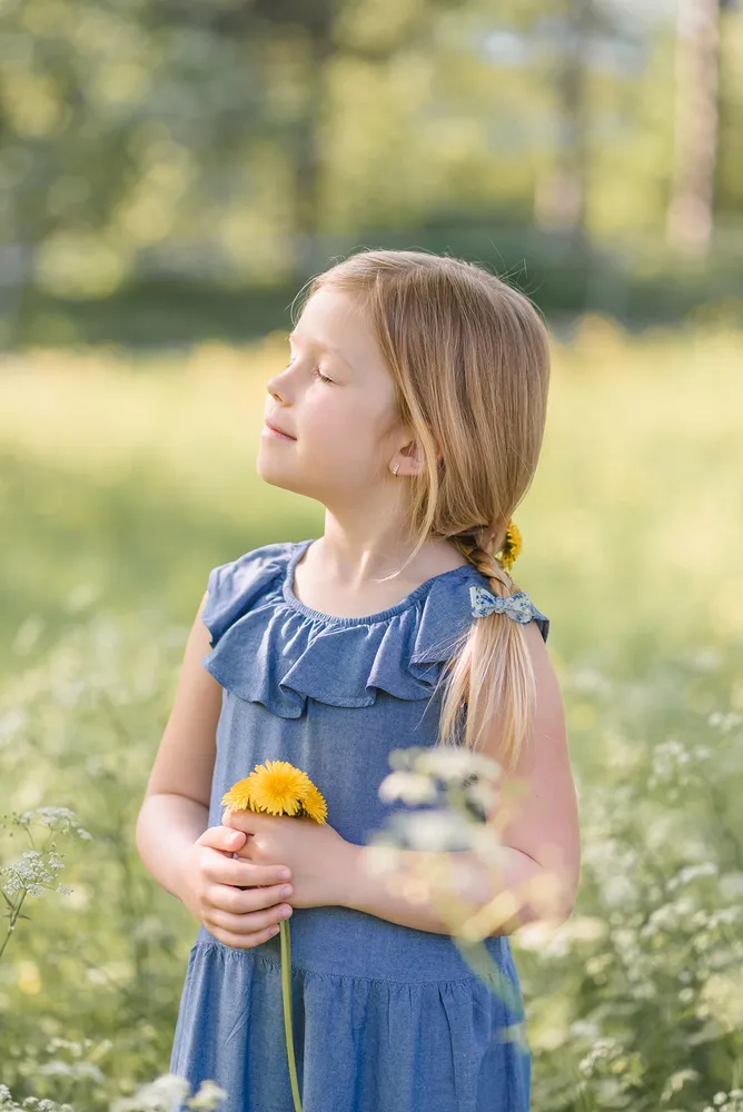 Girl in denim dress holds yellow flower, standing in sunlit meadow with trees in background, eyes closed, hair in braid with blue bow.