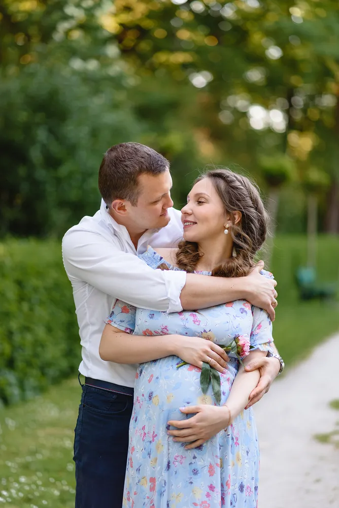 Expectant couple shares tender moment in a serene park setting, surrounded by lush greenery and soft sunlight. The man in a white shirt and dark pants embraces the woman, who wears a floral dress and gently cradles her belly. Their affectionate gaze and gentle touch convey warmth and anticipation. The blurred background highlights their intimate connection, capturing a peaceful and hopeful moment in nature.