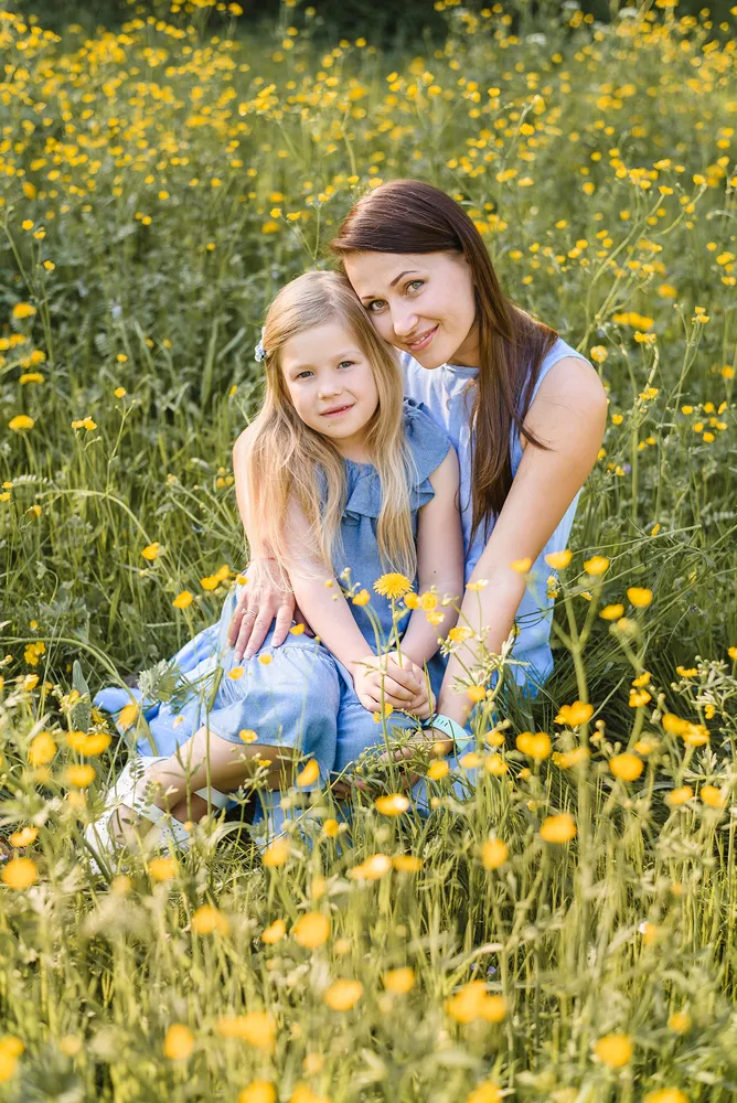 Woman and young girl in matching blue dresses sit closely in a field of tall yellow flowers, smiling softly at the camera. Natural lighting highlights their serene expressions and the vibrant surroundings.
