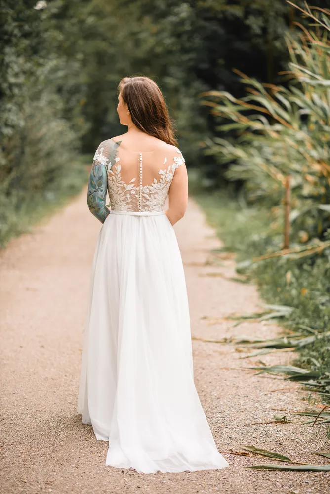 Woman in white lace-back wedding gown walks down gravel path, tattoo visible on right arm, surrounded by blurred greenery.
