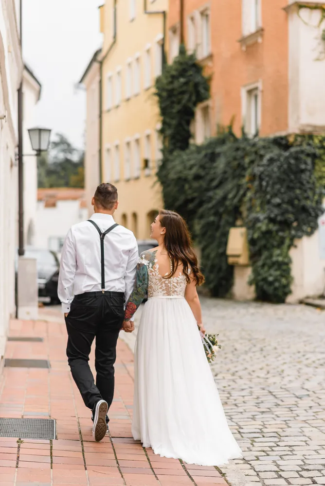 Bride and groom stroll hand-in-hand down a cobblestone street, surrounded by colorful European buildings, on their wedding day.
