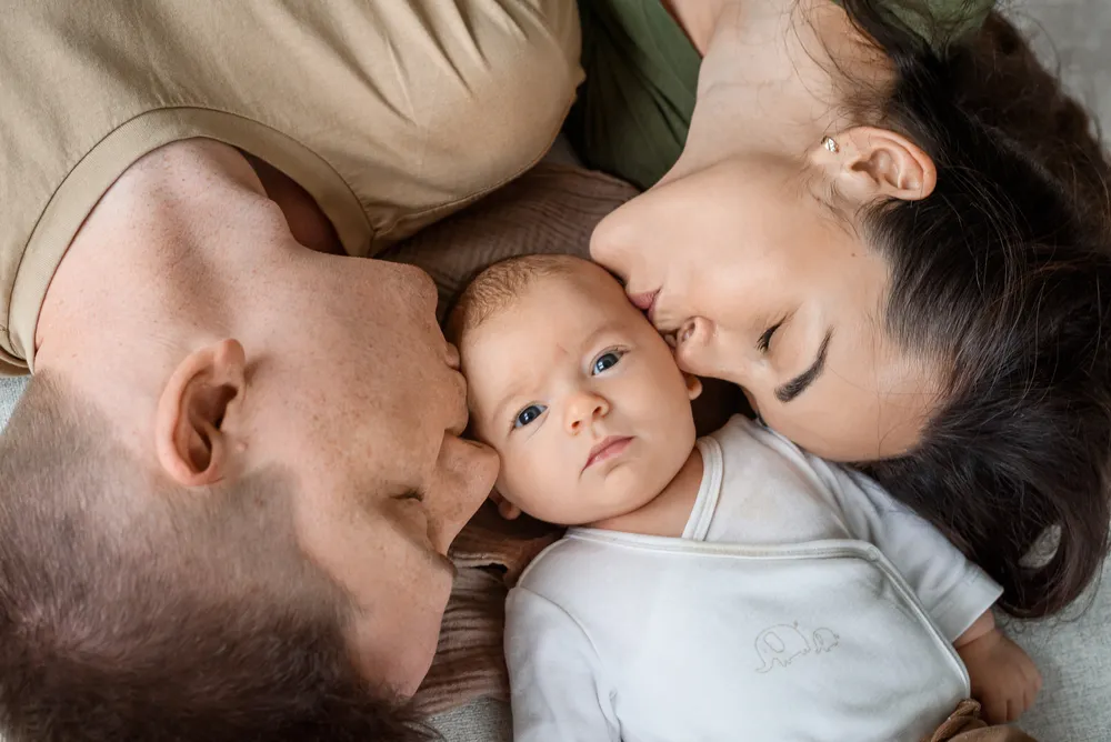 A newborn baby with blue eyes lies on a gray blanket, flanked by a man and woman in a tender family embrace. The man, with a closely shaved head, and the woman, with dark hair, kiss the baby's cheeks. The man wears a beige shirt, and the woman is in a green top. The baby is dressed in a white onesie with a subtle elephant design.