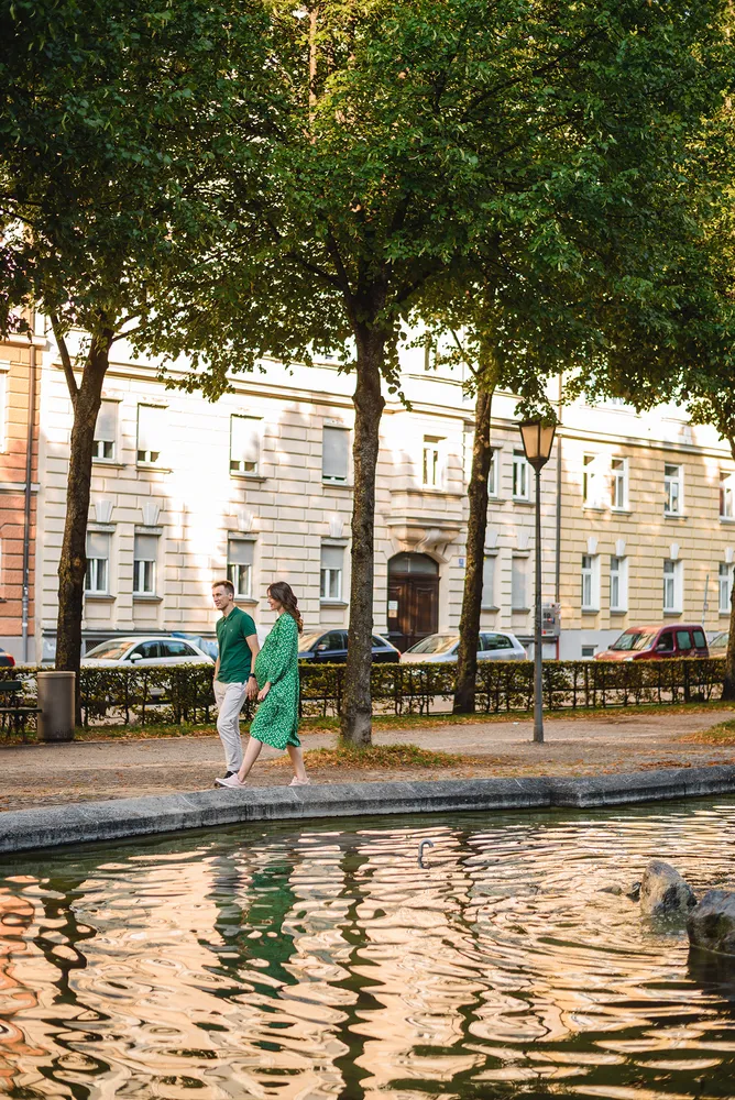 Pregnant woman in green dress walks with man along tree-lined canal in urban park setting, reflecting buildings and foliage on water.