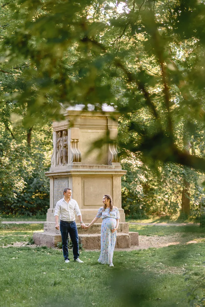 Couple in white shirts and floral dress holds hands in park, standing near stone monument with tree branches framing scene.