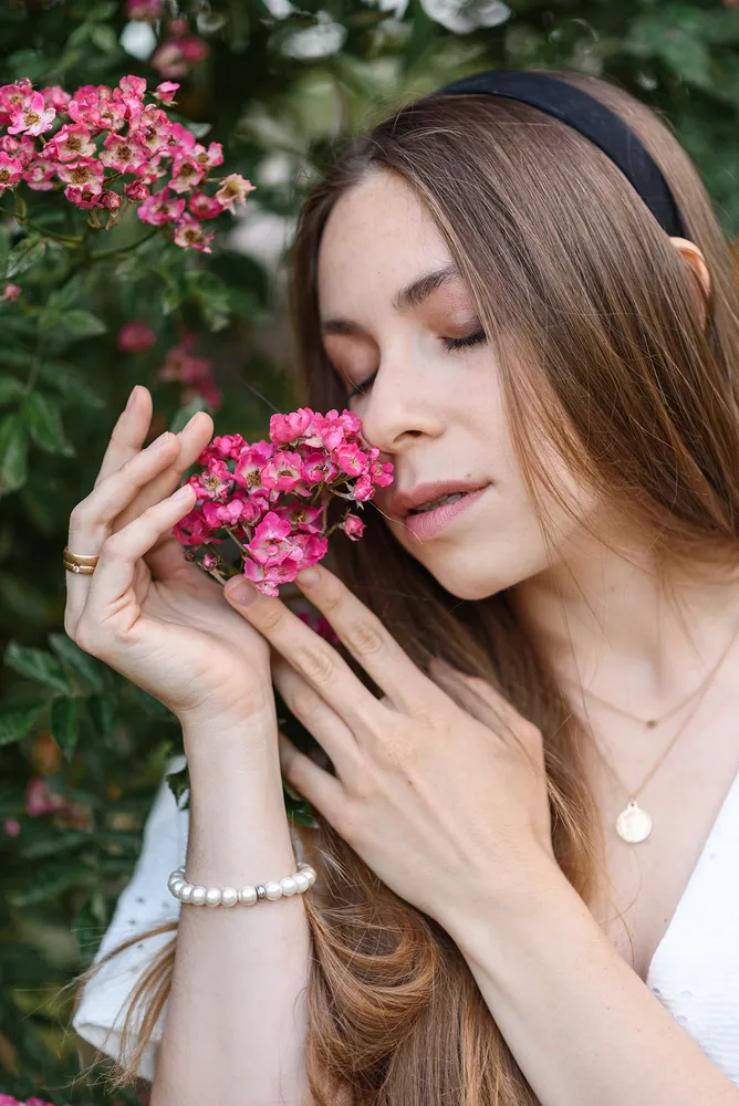 Woman with long brown hair, white top, and black headband holds pink flowers close to her face, standing in front of greenery. She wears a pearl bracelet and gold necklace.